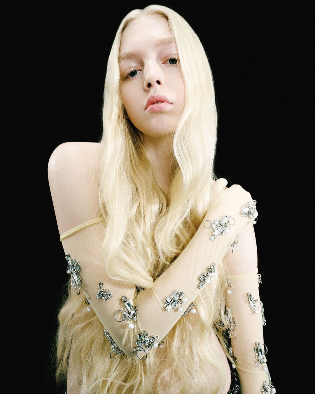 Portrait of a young woman with long blond hair and fair skin, wearing a sheer beaded top, posing against a solid black background.