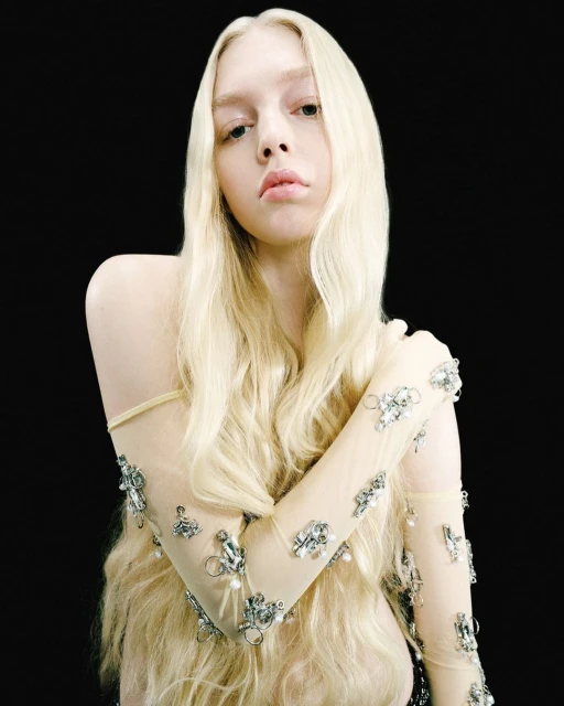 Portrait of a young woman with long blond hair and fair skin, wearing a sheer beaded top, posing against a solid black background.