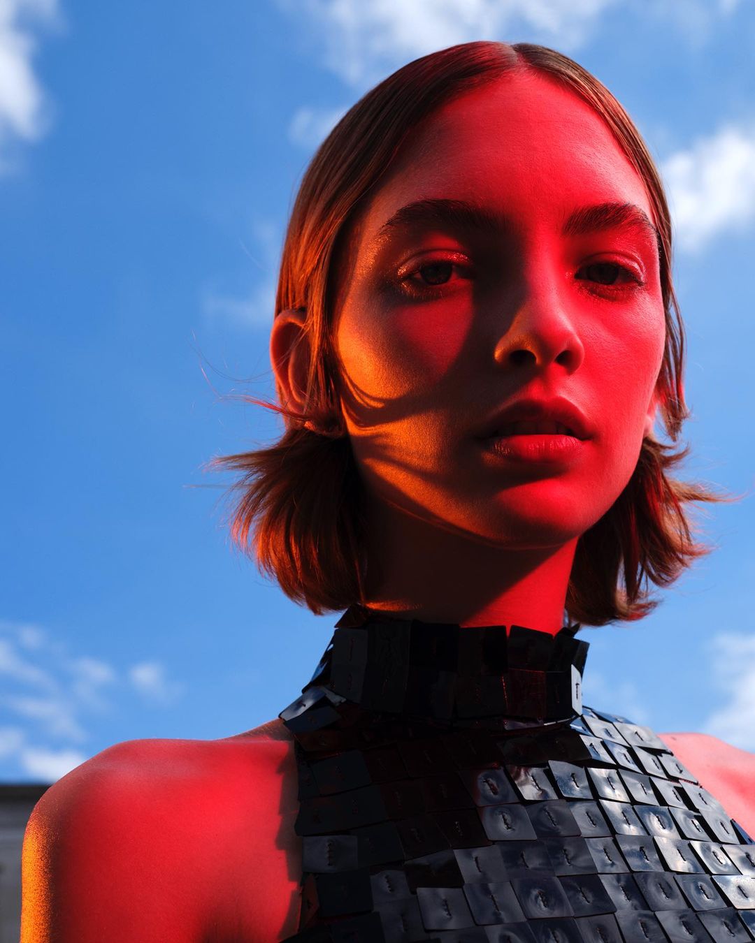 Portrait of an adult female with short hair wearing a textured dark top, posing against a bright blue sky with soft white clouds.
