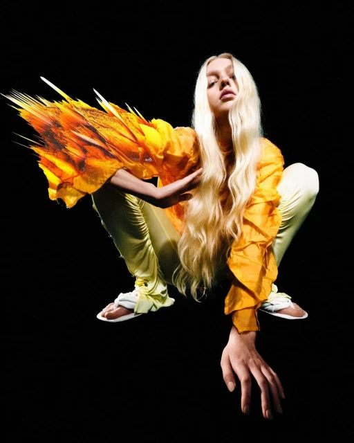 A woman with long blonde hair is crouched in a studio setting, wearing an orange ruffled top and light green pants against a black background.