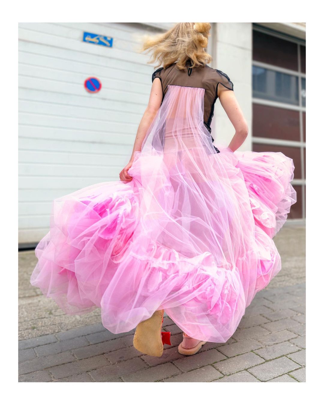 Adult female in motion wearing a black top and a large pink tulle gown outdoors in an urban environment in daylight, rear view.
