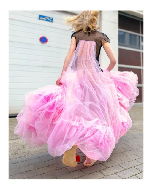 Adult female in motion wearing a black top and a large pink tulle gown outdoors in an urban environment in daylight, rear view.