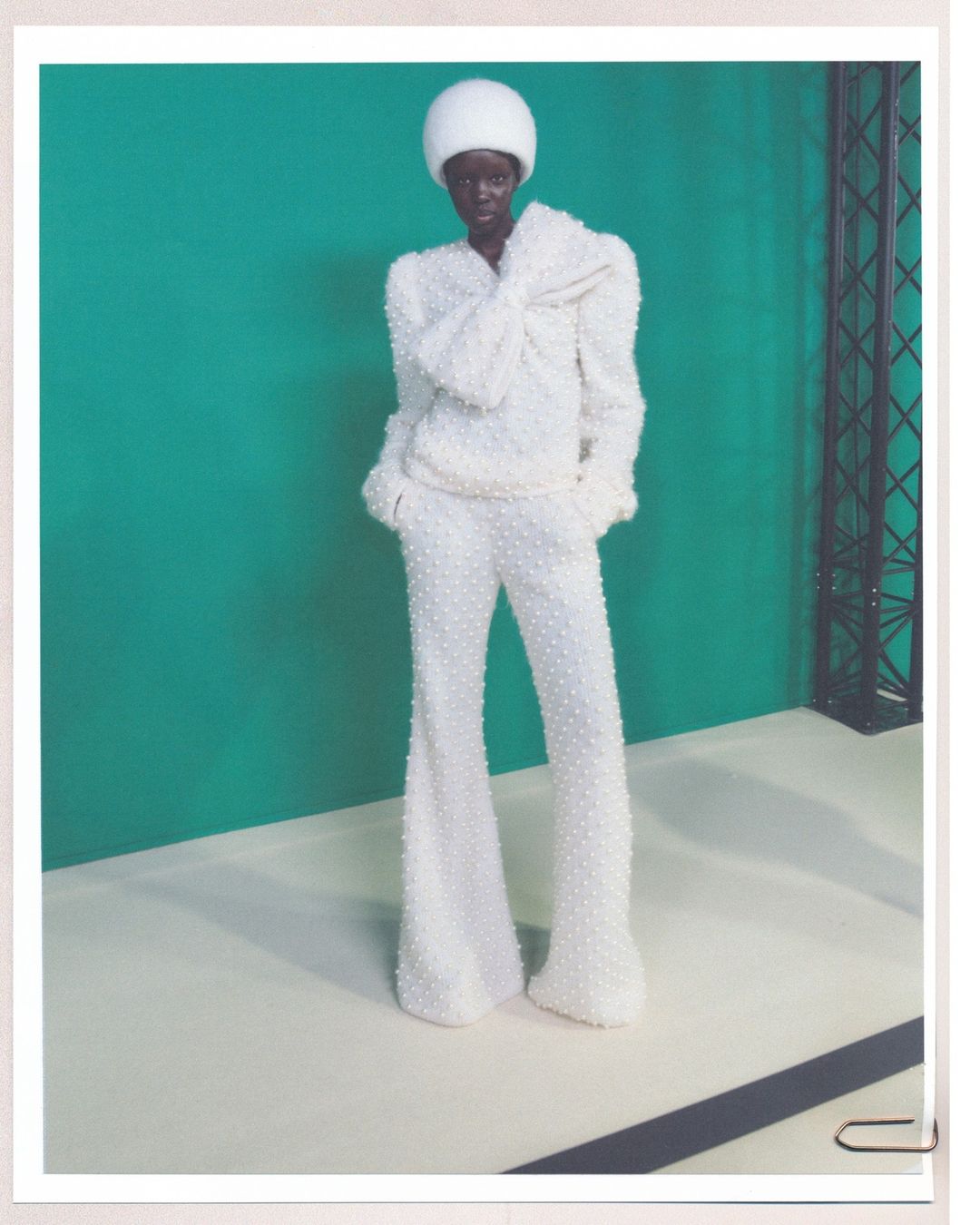Fashion shot of a dark-skinned woman in white beaded pantsuit and matching hat posing against a teal background. Full-length studio portrait.