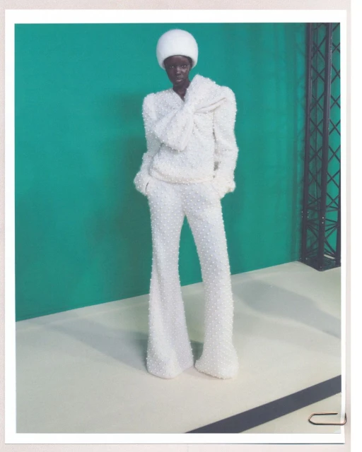 Fashion shot of a dark-skinned woman in white beaded pantsuit and matching hat posing against a teal background. Full-length studio portrait.