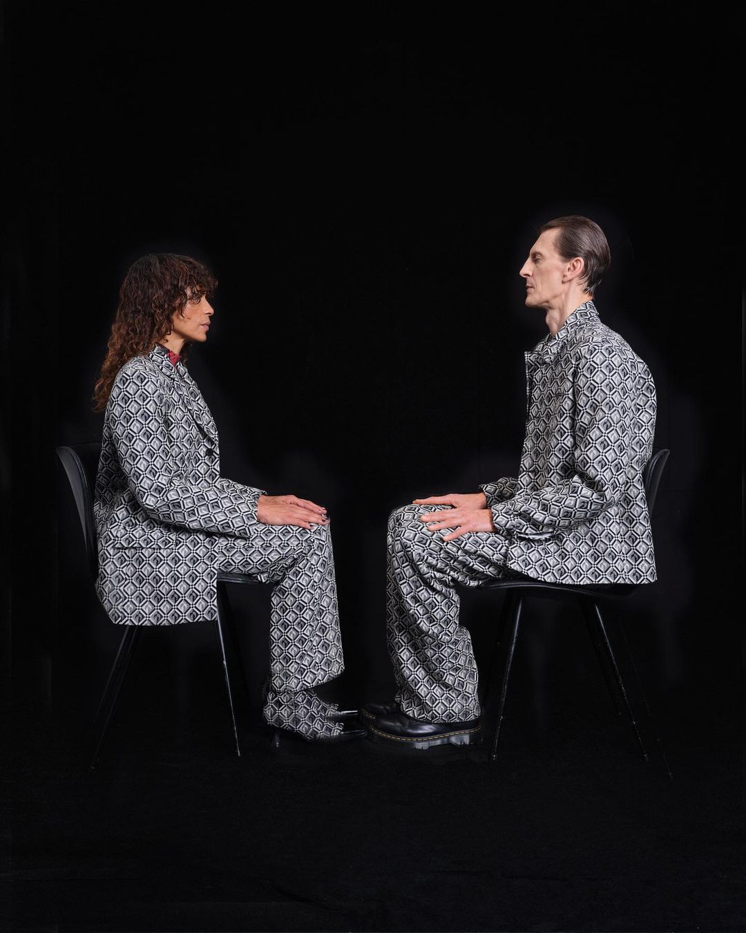 Two people in patterned suits sit facing each other in a minimalist setting. Black backdrop with studio lighting and matching suit sets.