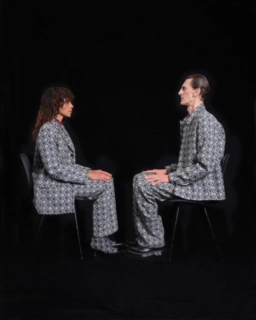 Two people in patterned suits sit facing each other in a minimalist setting. Black backdrop with studio lighting and matching suit sets.