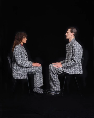 Two people in patterned suits sit facing each other in a minimalist setting. Black backdrop with studio lighting and matching suit sets.