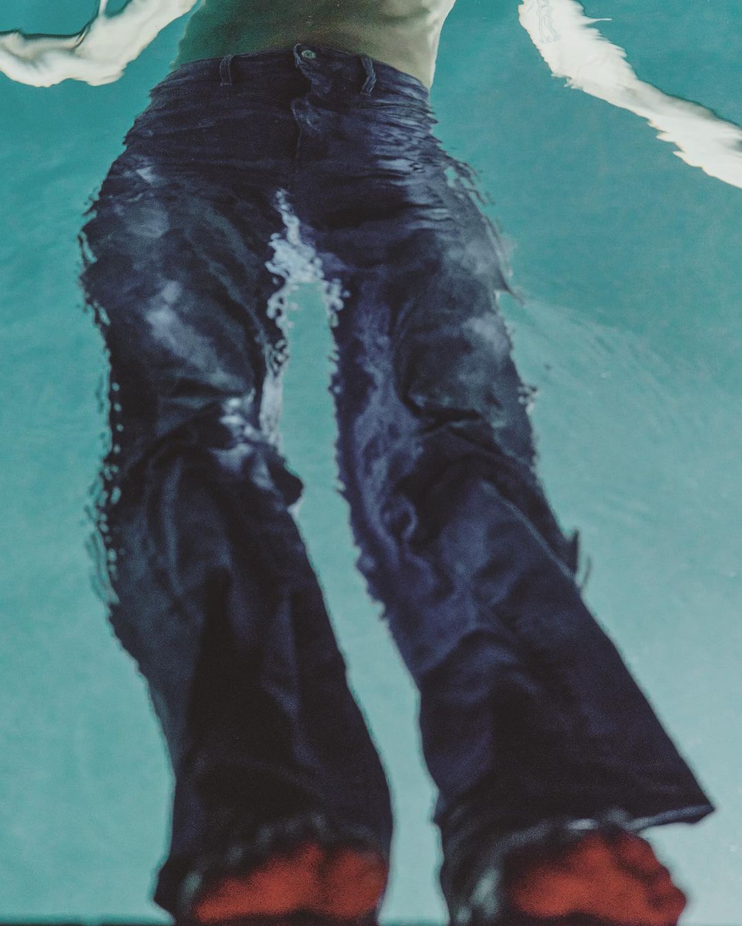 Person submerged underwater wearing dark denim jeans and red shoes, creating a surreal and artistic fashion image.