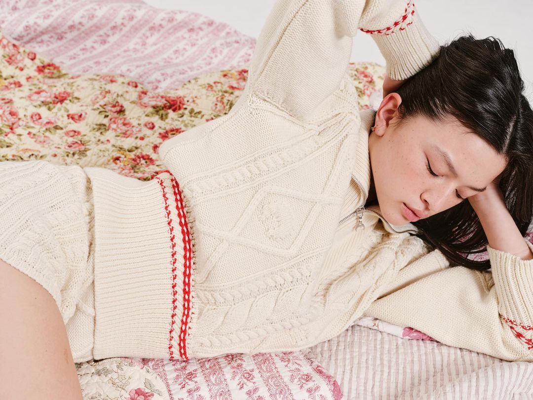 Young woman with dark hair relaxes on a bed featuring floral patterned bedding, wearing a cream-colored knit top with red accents.