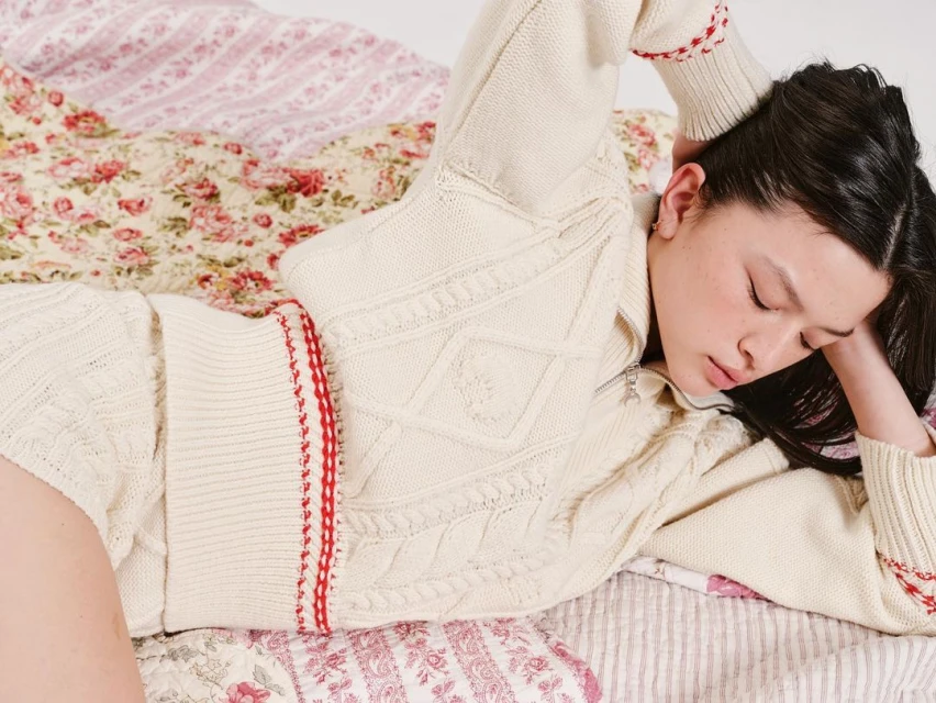 Young woman with dark hair relaxes on a bed featuring floral patterned bedding, wearing a cream-colored knit top with red accents.