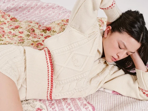 Young woman with dark hair relaxes on a bed featuring floral patterned bedding, wearing a cream-colored knit top with red accents.