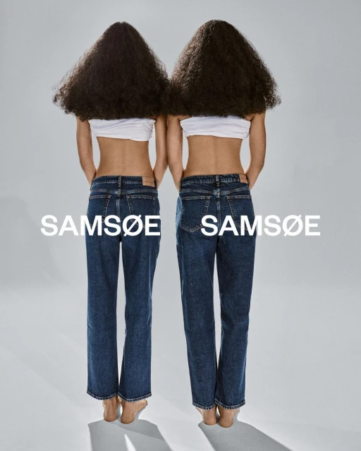 Back view of two women wearing blue jeans and white tops, standing in a bright studio against a solid grey backdrop. Fashion shoot.