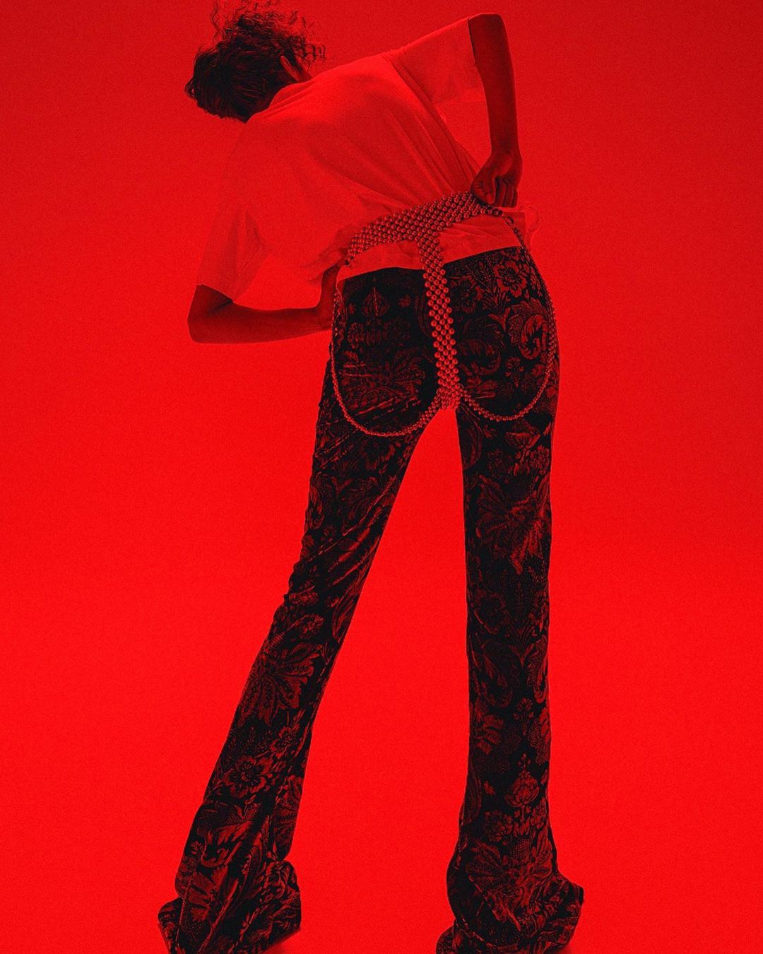 Back view of a person wearing a white shirt, patterned pants, and a beaded belt against a solid red background in a studio setting.