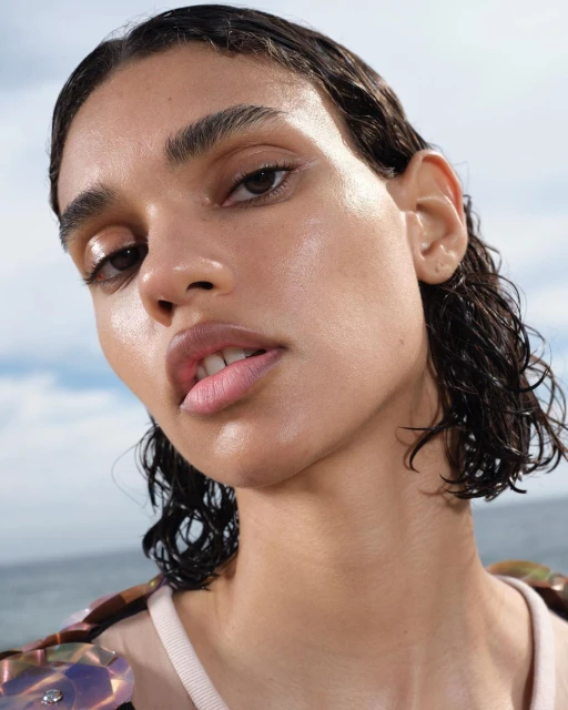 Close-up portrait of a woman with wet, short hair and natural makeup against a sky and water background. Beauty and skincare focus.