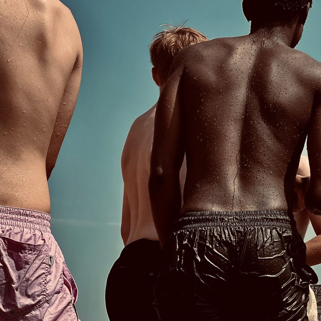 Three shirtless men in swimwear standing against a light blue background. One man is fair-skinned and two are dark-skinned.