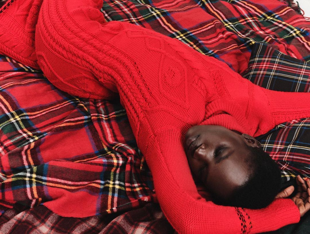 Woman in a red cable-knit jumpsuit reclines on a red plaid blanket, eyes closed, showcasing a cozy and comfortable fashion aesthetic.
