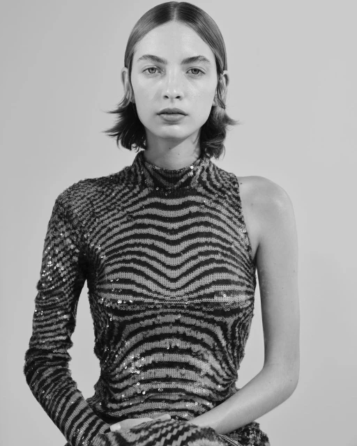 Black and white portrait of a young woman with short hair wearing a one-shoulder, zebra-striped, sequined dress, gazing intently at the camera.