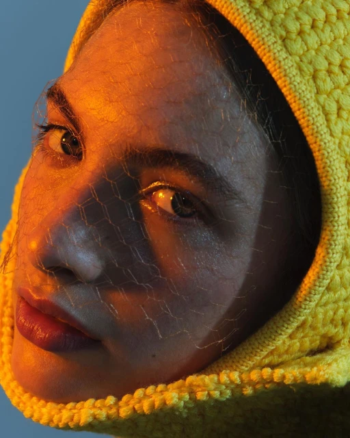 Close-up portrait of an adult female with fair skin wearing a yellow knitted hood and sheer veil, set against a blue background.