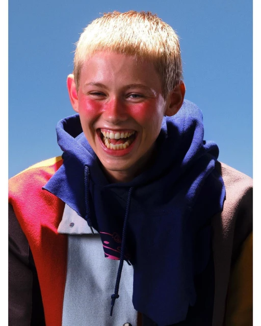 A young adult with blond hair grins widely in a close-up shot. The individual wears a hoodie and jacket against a clear blue sky.