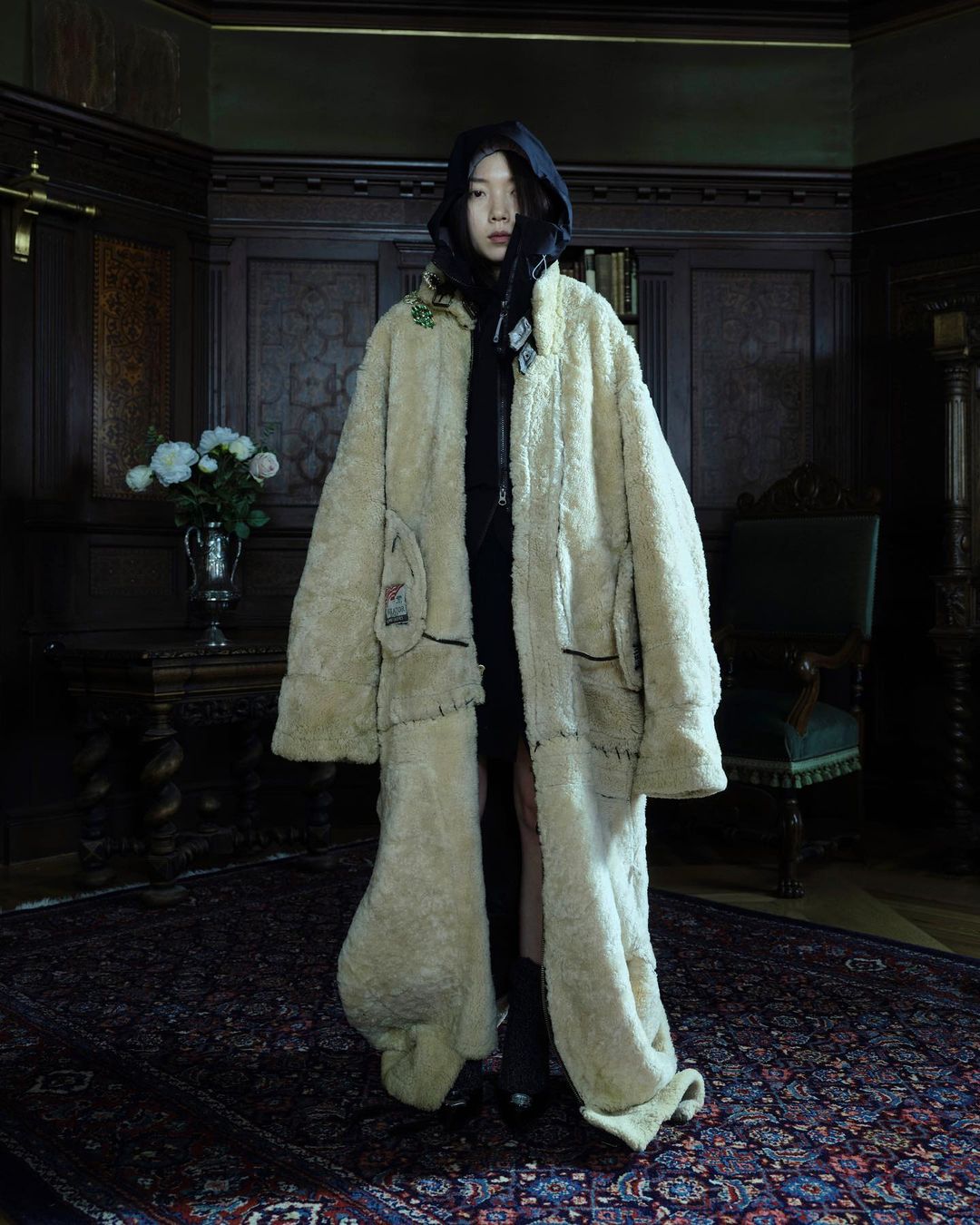 Asian woman in oversized faux fur coat poses in interior with dark wood panelling, patterned rug, chair, table with flowers, and dark lighting.