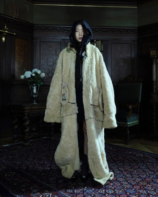 Asian woman in oversized faux fur coat poses in interior with dark wood panelling, patterned rug, chair, table with flowers, and dark lighting.