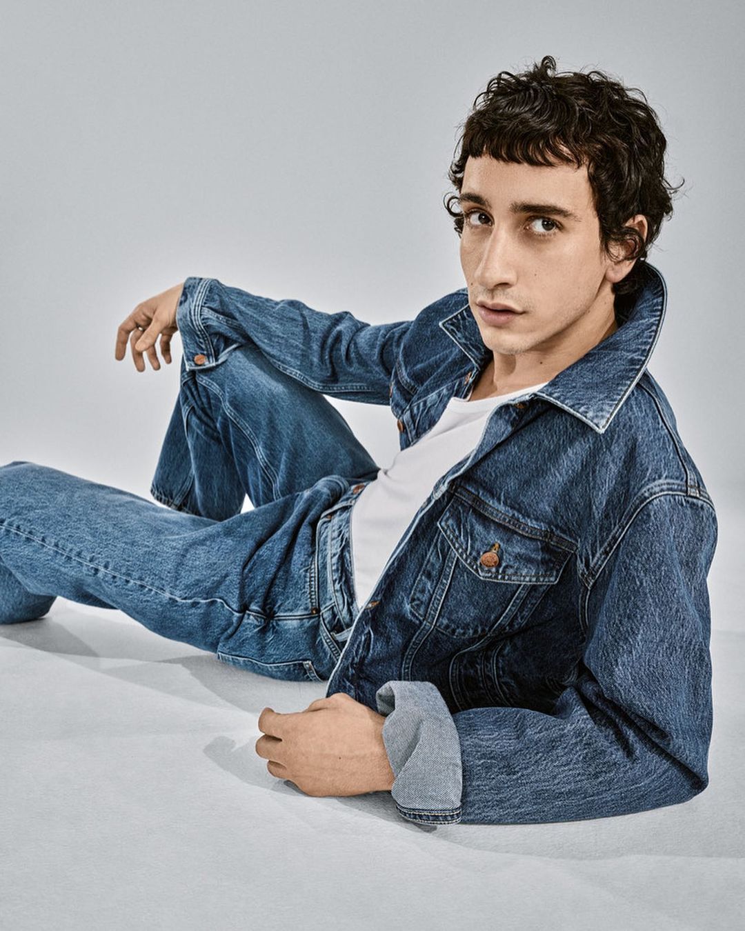 A young male model with dark curly hair wearing a denim jacket and jeans poses on a plain white studio backdrop.