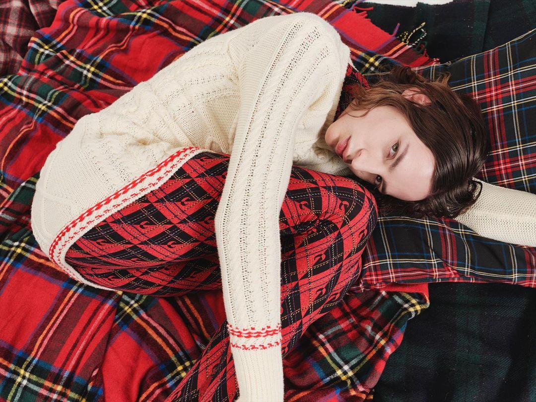 A young adult poses in a cream-colored sweater and red plaid pants on a plaid blanket in a studio setting for a fashion shot.