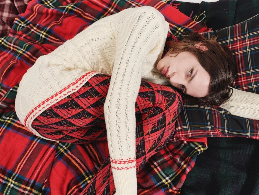 A young adult poses in a cream-colored sweater and red plaid pants on a plaid blanket in a studio setting for a fashion shot.