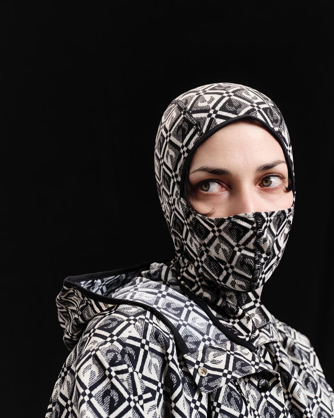 Portrait of a woman wearing a patterned balaclava-style hood and jacket against a black background, looking off to the side.