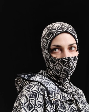 Portrait of a woman wearing a patterned balaclava-style hood and jacket against a black background, looking off to the side.