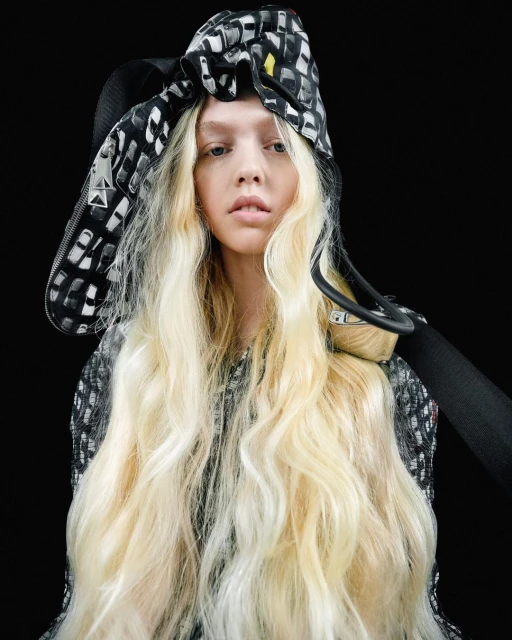 Fashion portrait of a young woman with long blonde hair wearing a patterned headscarf and matching top against a dark background.