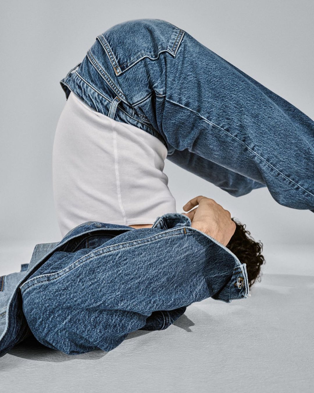 Person in blue denim jacket and jeans poses with bent knees and a white tee shirt against a grey backdrop in a fashion shoot.