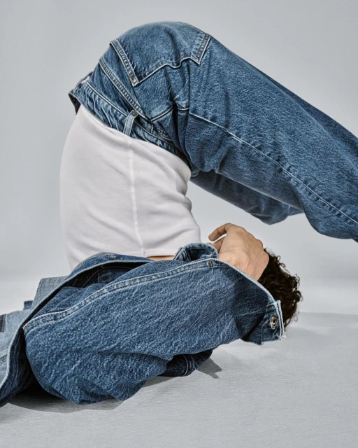 Person in blue denim jacket and jeans poses with bent knees and a white tee shirt against a grey backdrop in a fashion shoot.