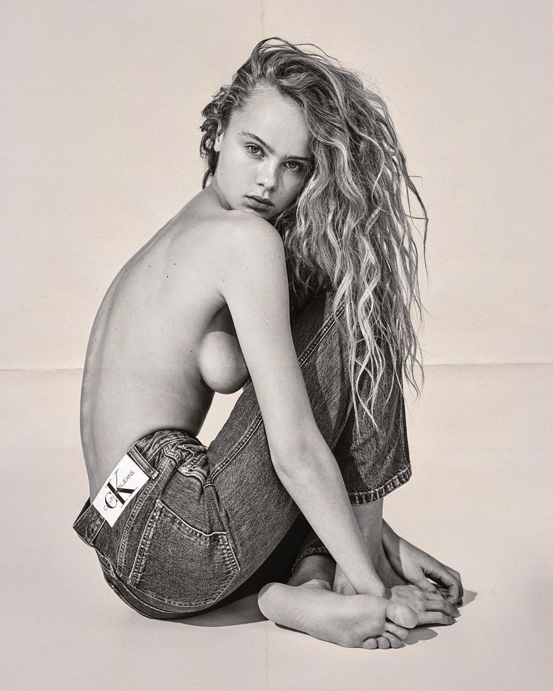 Monochrome portrait of a bare-chested woman wearing jeans, seated with her knees up, showcasing wavy hair against a neutral backdrop.