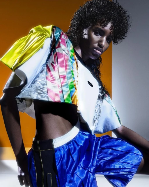 Model poses in a vibrant fashion shot featuring a colorful jacket with yellow, white, and floral panels and blue shorts against a geometric backdrop.