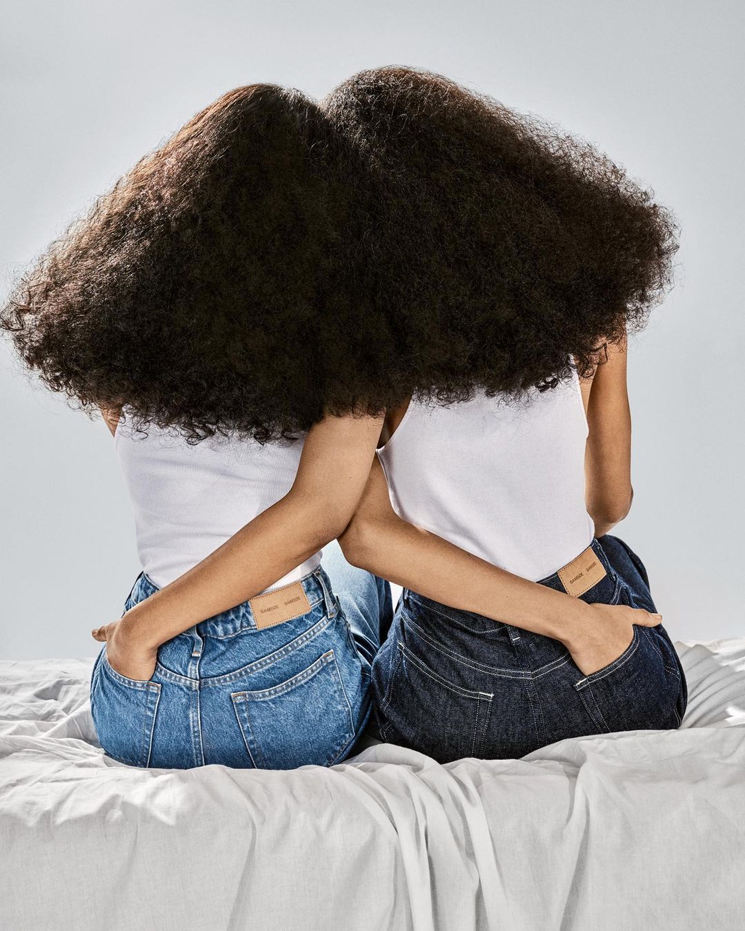 Two women with large dark curly hair sit close together on a white sheet wearing jeans and tank tops in a brightly lit photo studio.