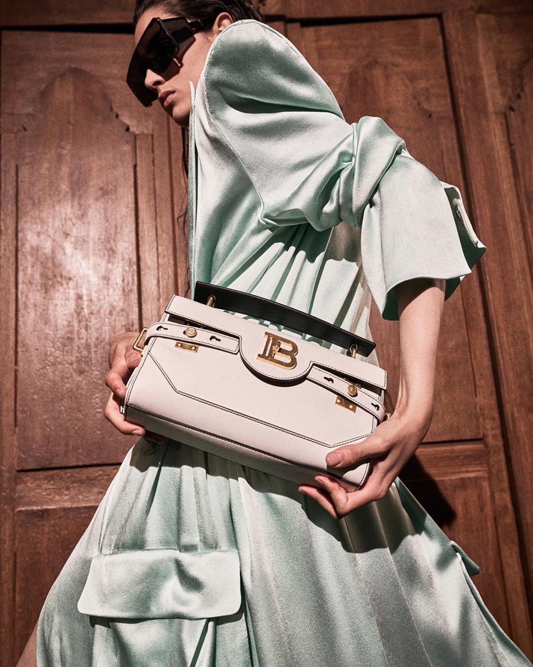 An adult female poses indoors in a light green dress with sunglasses, holding a Balmain handbag with a wood door background.