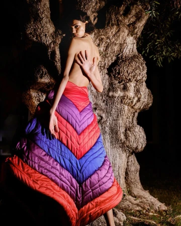 Model with bare back wearing a voluminous quilted chevron skirt in red, purple, and blue standing against a large textured tree trunk.
