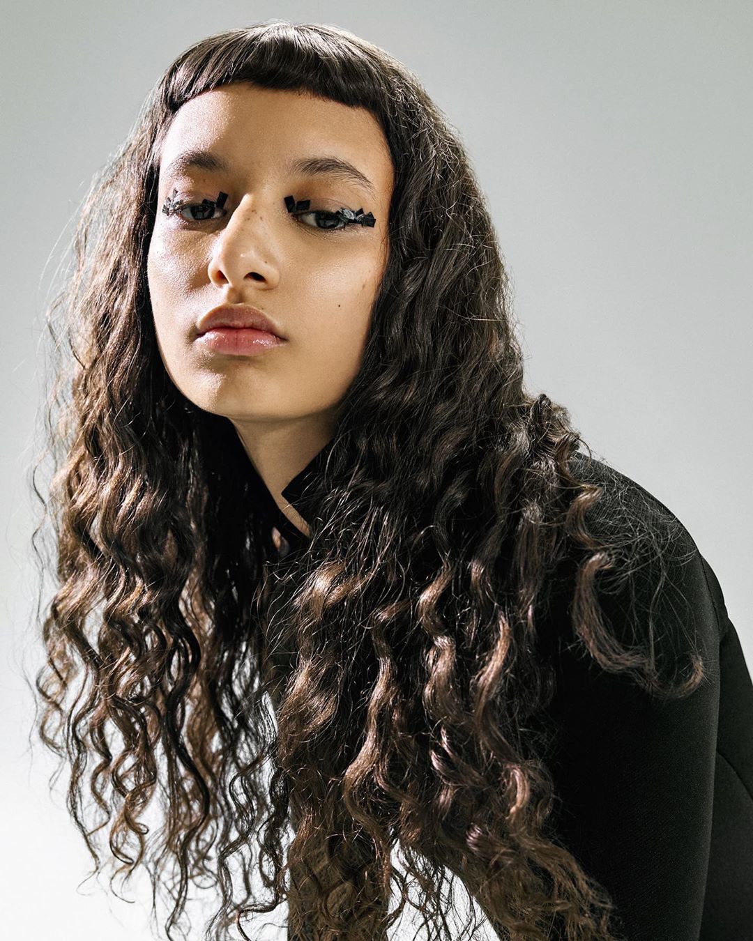 Close-up portrait of a woman with curly hair and artistic black eye makeup against a gradient gray backdrop in a beauty fashion shot.
