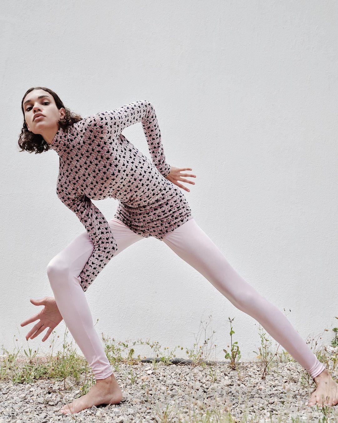 Model in printed top and pale pink leggings posing outdoors on gravel against a textured white wall.