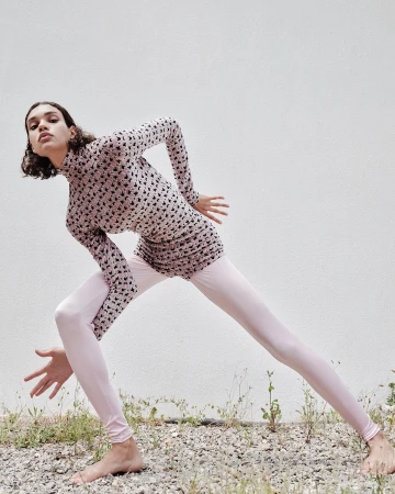 Model in printed top and pale pink leggings posing outdoors on gravel against a textured white wall.