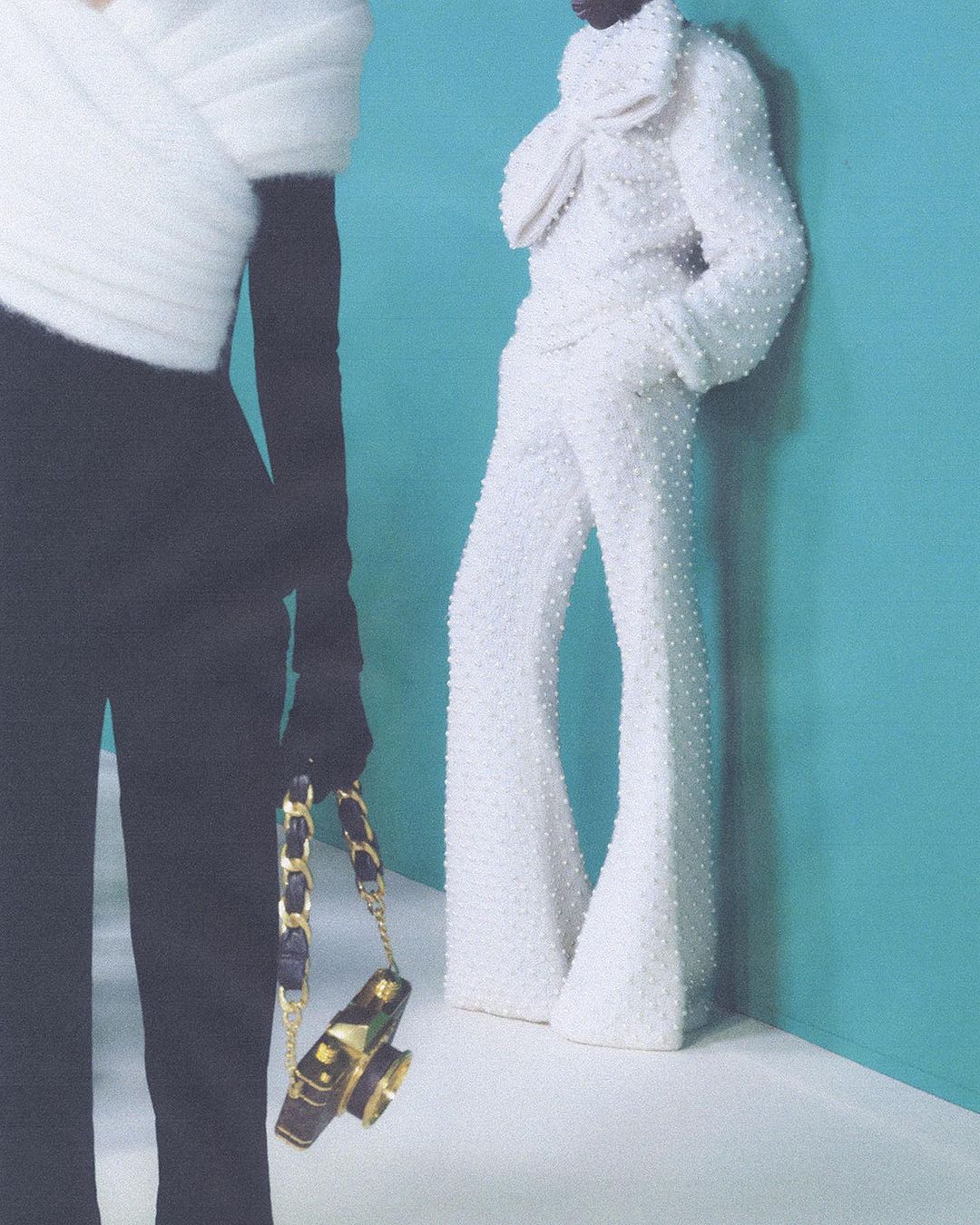 Two fashion models pose indoors against a turquoise backdrop. One wears a black top and pants, the other an all-white beaded outfit.