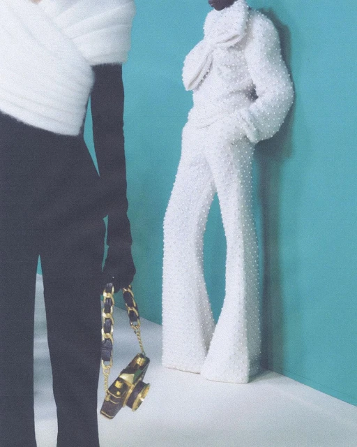 Two fashion models pose indoors against a turquoise backdrop. One wears a black top and pants, the other an all-white beaded outfit.