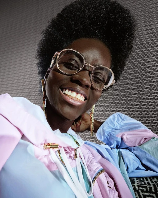 Close-up shot of a smiling, dark-skinned woman wearing glasses and a colourful pastel jacket in front of a patterned wall.