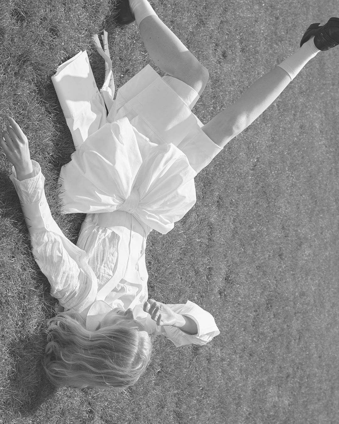 Monochrome image of a person in white clothing lying on green grass, legs in the air, with soft lighting, and an unusual angle.