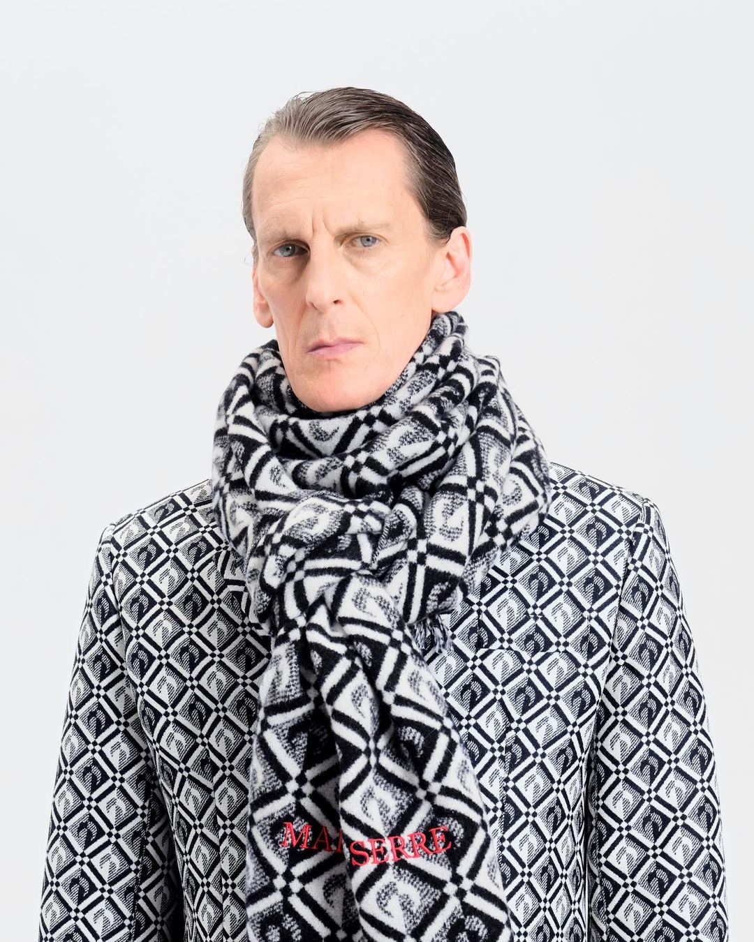 A fair-skinned adult male poses indoors wearing a black-and-white patterned jacket and scarf against a bright white backdrop.