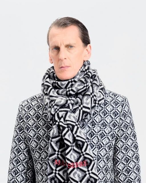 A fair-skinned adult male poses indoors wearing a black-and-white patterned jacket and scarf against a bright white backdrop.