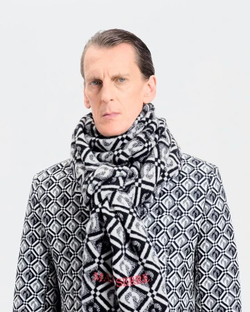 A fair-skinned adult male poses indoors wearing a black-and-white patterned jacket and scarf against a bright white backdrop.