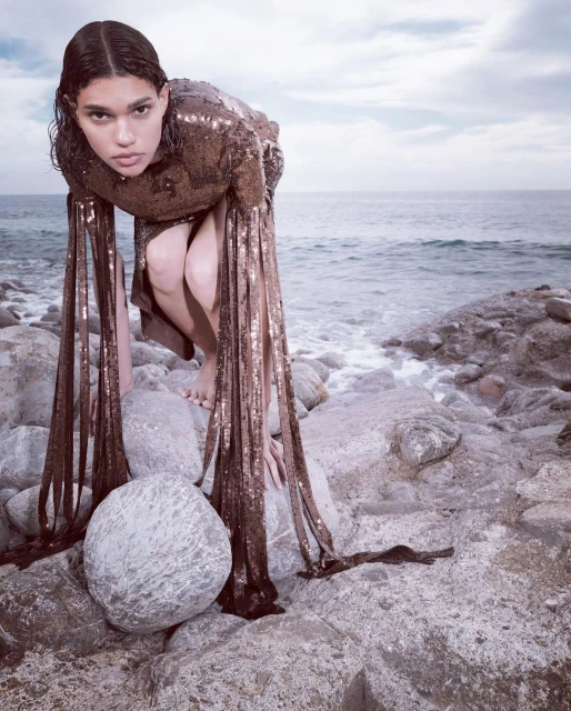 Woman in shimmering dress poses on rocks at the coast. Ocean view with a bright, overcast sky. Fashion editorial shot.