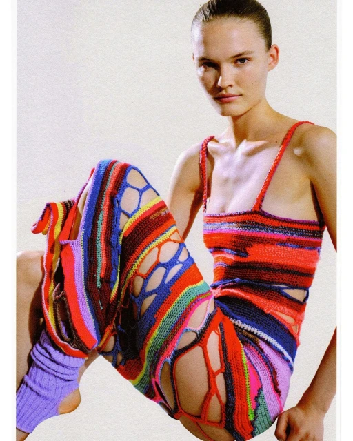 A model poses in a colorful knitted top and pants with cutouts against an off-white backdrop. The shot is a medium portrait, emphasizing the fashion.
